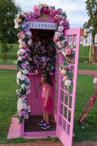 A young girl plays in a flower-filled pink telephone booth in a park.
