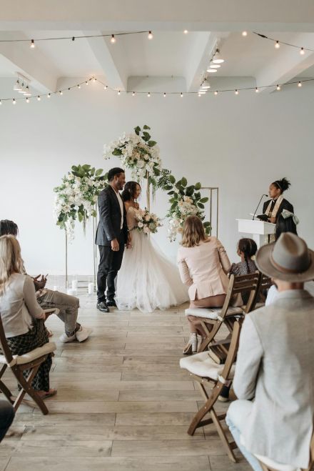 A small gathering witnesses a beautiful wedding ceremony indoors with wooden chairs and floral decor.